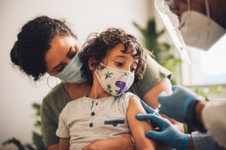 Little boy wearing face mask taking vaccine at home. Kid with mother receiving covid vaccine from a healthcare worker at home.